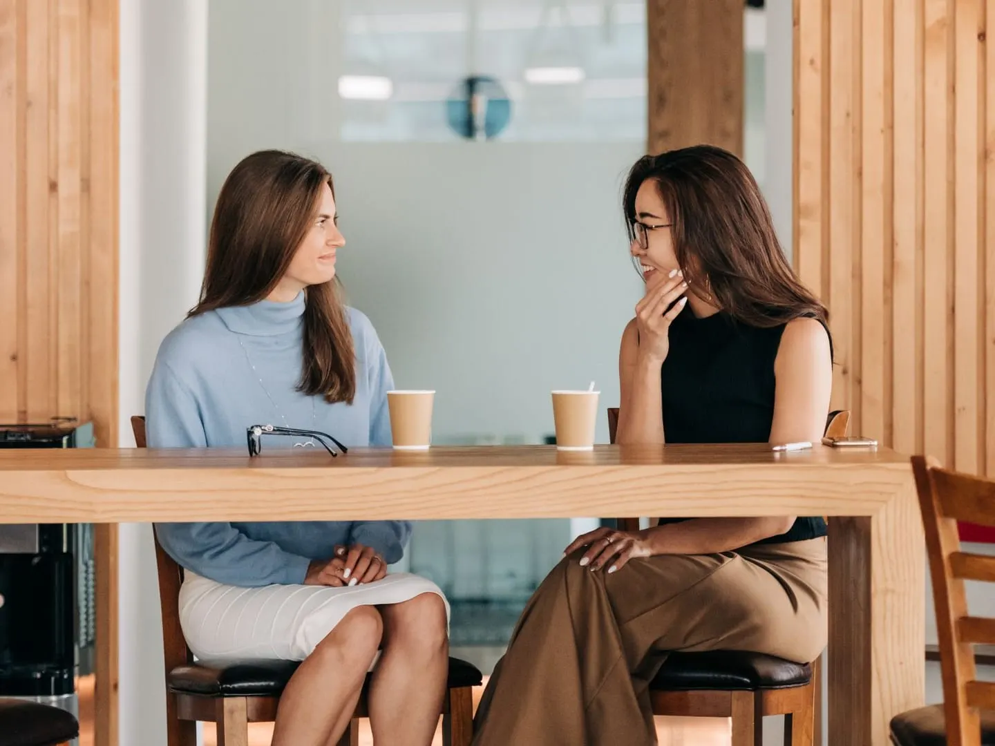 Two women in casual clothes chatting and smiling over coffee at a wooden table