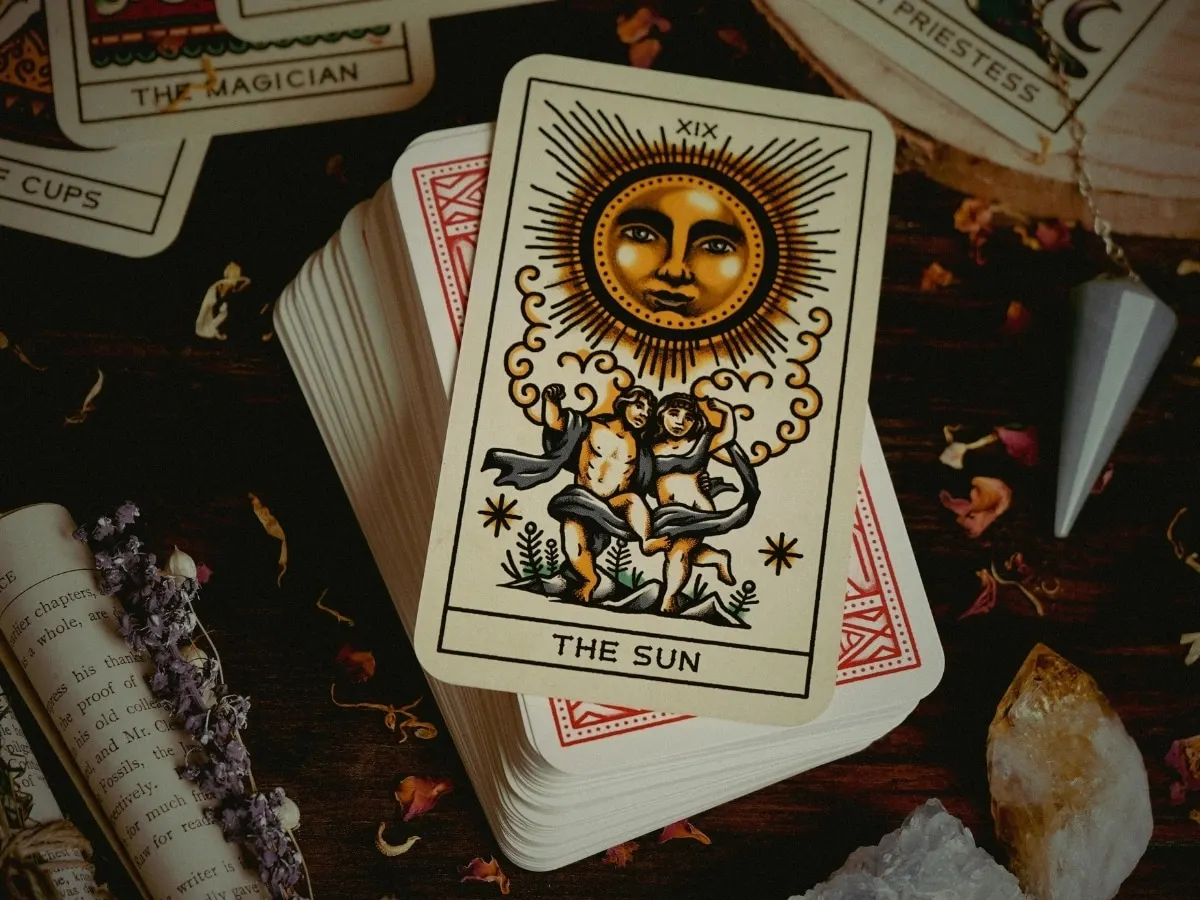 A tarot deck fanned on a dark wooden table, The Sun card on top, with crystals and dried flowers